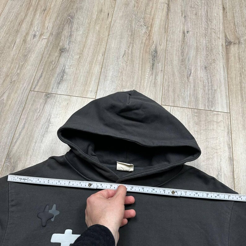 Może przedstawiać: Ciemnoszara bluza z kapturem z kapturem i białą metką. Bluza z kapturem ma biało-szary wz&oacute;r z przodu. Linijka jest trzymana na bluzie z kapturem, aby zmierzyć jej szerokość.