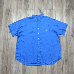 Puede incluir: Camisa azul brillante de manga corta con botones. La camisa tiene un cuello clásico y un pequeño logotipo amarillo en el pecho. La camisa está hecha de una tela ligera y tiene un ajuste relajado. La camisa está colocada sobre una superficie de madera.