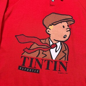 Pu&ograve; includere: Maglietta rossa a maniche lunghe con un'illustrazione a cartoni animati di Tintin, un reporter, con un abito e un berretto marroni e una cravatta rossa. La parola "TINTIN" &egrave; stampata in nero, con "REPORTER" sotto. Colletto abbottonato. &copy; Herg&eacute;, 1994.