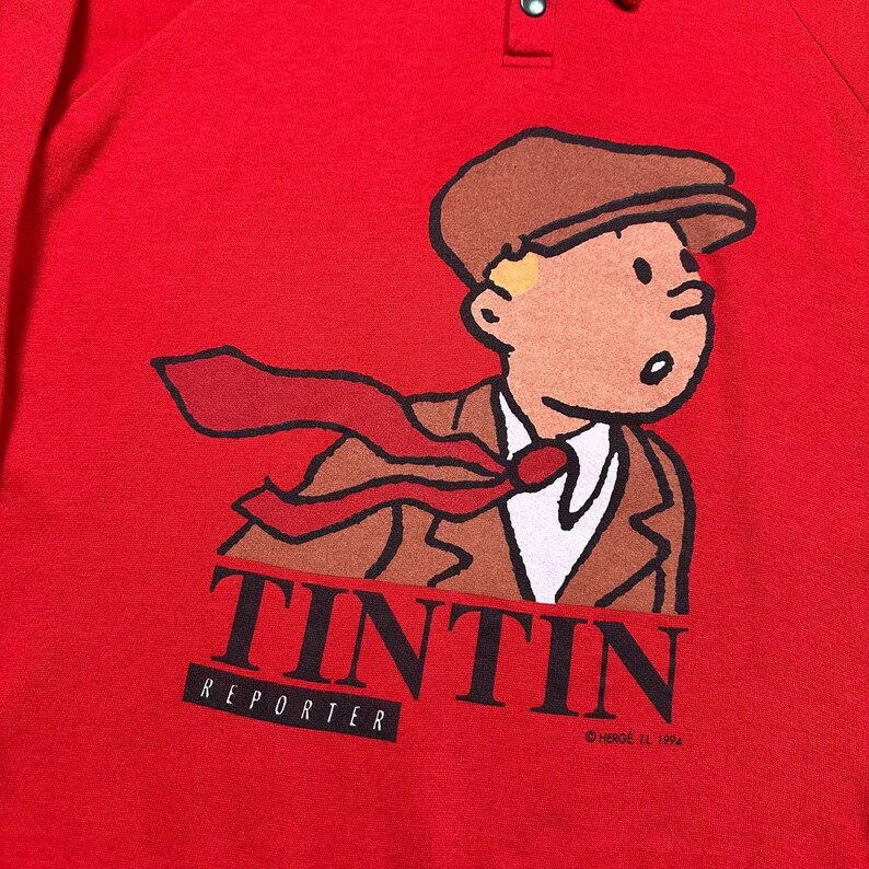 Pu&ograve; includere: Maglietta rossa a maniche lunghe con un'illustrazione a cartoni animati di Tintin, un reporter, con un abito e un berretto marroni e una cravatta rossa. La parola "TINTIN" &egrave; stampata in nero, con "REPORTER" sotto. Colletto abbottonato. &copy; Herg&eacute;, 1994.