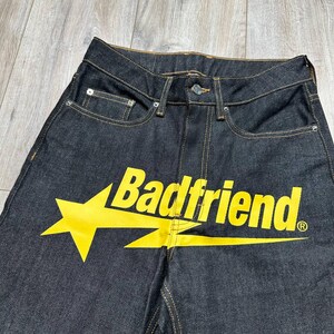 May include: Dark wash denim jeans with a yellow star and the text "Badfriend" printed across the front. The jeans have classic five-pocket styling and a button and zipper closure.