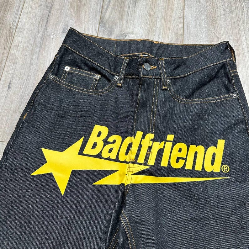 May include: Dark wash denim jeans with a yellow star and the text "Badfriend" printed across the front. The jeans have classic five-pocket styling and a button and zipper closure.