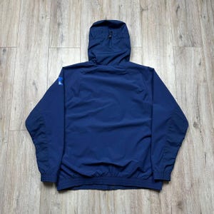 Może przedstawiać: Granatowa kurtka z kapturem, zapinana na zamek błyskawiczny, ze ściągaczami w mankietach i u dołu. Kurtka ma małe logo na lewym rękawie. Kurtka wykonana jest z lekkiego, wodoodpornego materiału.