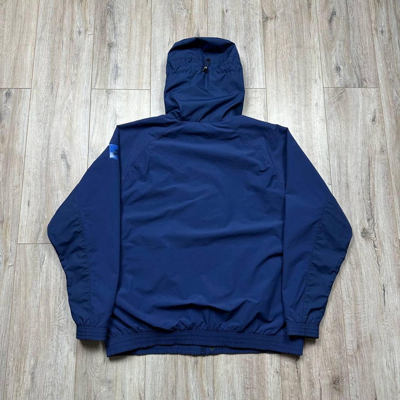 Może przedstawiać: Granatowa kurtka z kapturem, zapinana na zamek błyskawiczny, ze ściągaczami w mankietach i u dołu. Kurtka ma małe logo na lewym rękawie. Kurtka wykonana jest z lekkiego, wodoodpornego materiału.