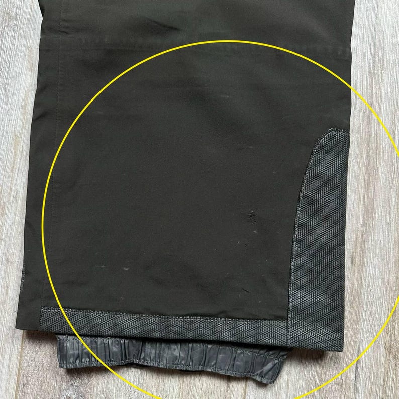 May include: Close-up of black pants with a textured, reinforced patch on the knee area. The pants have a dark, matte finish and a gathered, elasticized hem. The fabric appears to be a durable, weather-resistant material.