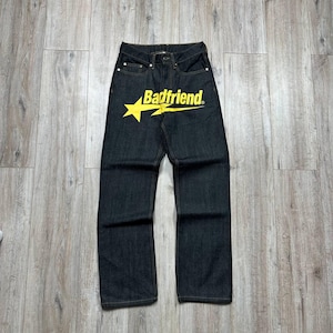 May include: Dark wash denim jeans with a straight leg cut. The jeans feature a yellow star graphic with the text "Badfriend" across the front. The jeans have classic five-pocket styling.