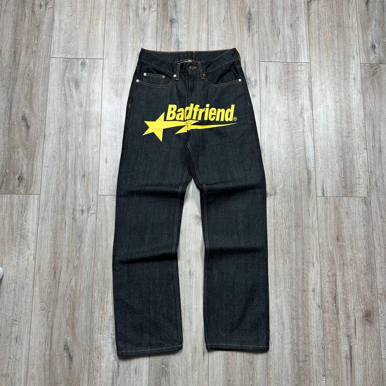 May include: Dark wash denim jeans with a straight leg cut. The jeans feature a yellow star graphic with the text "Badfriend" across the front. The jeans have classic five-pocket styling.