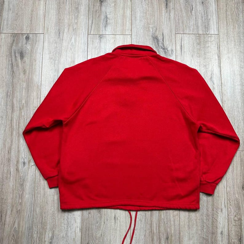 Pu&ograve; includere: Giacca sportiva rosso brillante con collo alto e maniche lunghe. La giacca ha un cordoncino sull'orlo inferiore e maniche a raglan. Vista posteriore della giacca su una superficie di legno chiaro.