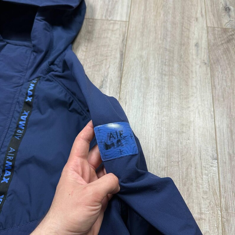 Może przedstawiać: Granatowa kurtka z kapturem. Kurtka ma nadruk "AIR MAX" w kolorze czarnym i niebieskim wzdłuż boku. Niebieska naszywka z napisem "AIR MAX" znajduje się na rękawie.