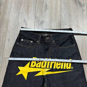 May include: Dark wash denim jeans with a yellow lightning bolt and the text "Badfriend". The jeans have a classic five-pocket design with a button and zipper closure. The jeans are being measured with a ruler.