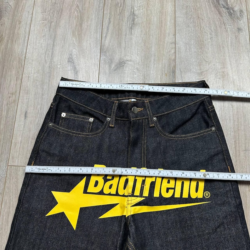 May include: Dark wash denim jeans with a yellow lightning bolt and the text "Badfriend". The jeans have a classic five-pocket design with a button and zipper closure. The jeans are being measured with a ruler.