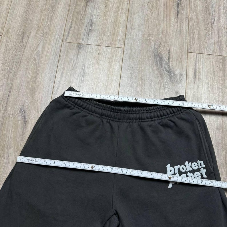 Może przedstawiać: Ciemnoszare spodnie dresowe z napisem "broken planet" w białym skrypcie na prawym dolnym udzie. Talia jest elastyczna. Taśma miernicza jest ułożona na g&oacute;rze i z boku spodni.