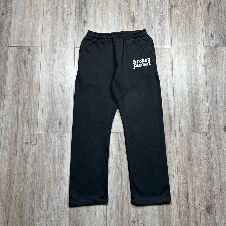 Może przedstawiać: Czarne spodnie dresowe z białym napisem "broken planet" na lewej g&oacute;rnej nogawce. Spodnie mają elastyczny pas i luźny kr&oacute;j. Materiał wydaje się miękki i wygodny.