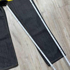 May include: Dark wash denim jeans with yellow stitching are measured with a white measuring tape. The jeans are laid flat on a light wood surface. The jeans are a straight leg style.