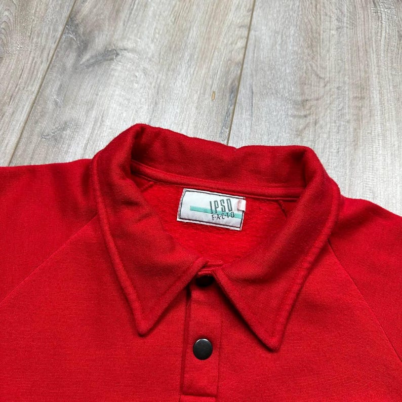Pu&ograve; includere: Primo piano di una polo rosso brillante con colletto classico e paramontura a due bottoni. La maglietta presenta il testo "IPSO FACTO" su un'etichetta vicino al colletto. Il tessuto sembra essere un materiale morbido a maglia.