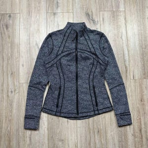 May include: A gray and black zip-up jacket with a high collar. The jacket has a textured pattern and black trim along the seams, zipper, and pockets. The jacket has long sleeves and a fitted silhouette.