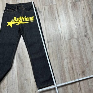 May include: Dark wash denim jeans with a yellow star and the text "Badfriend" printed on the front. The jeans are straight-legged and appear to be new. A measuring tape is placed next to the jeans.