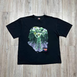 May include: Black t-shirt featuring a graphic of green aliens. The design includes a central alien figure with a glowing chest, surrounded by a group of similar figures. The shirt has a crew neck and short sleeves. The graphic has the text "Round FX".