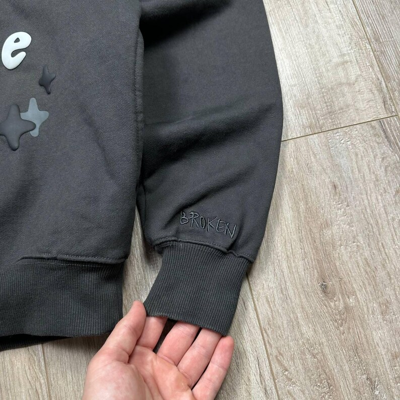Może przedstawiać: Ciemnoszara bluza z napisem "Broken" wyhaftowanym na mankiecie. Bluza posiada białą literę "e" i zdobienia w kształcie gwiazd na g&oacute;rze po lewej stronie. Tkanina wydaje się miękka i wygodna.