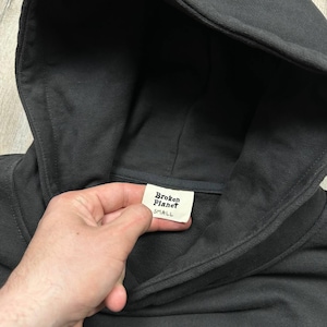 Może przedstawiać: Ciemnoszara bluza z kapturem z kapturem i metką z napisem "Broken Planet SMALL". Bluza z kapturem jest wykonana z miękkiego materiału i ma prosty, minimalistyczny design.