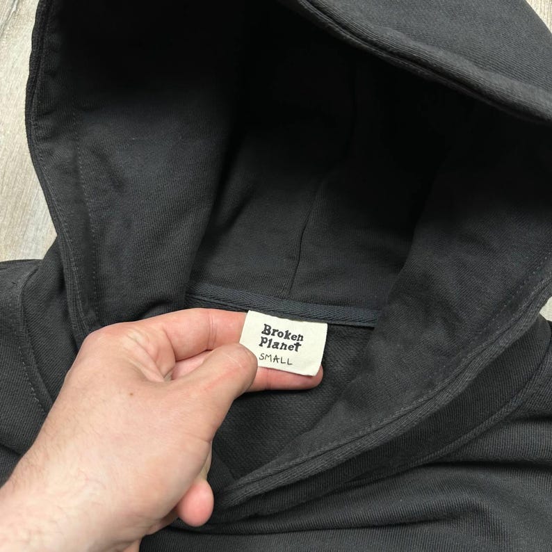 Może przedstawiać: Ciemnoszara bluza z kapturem z kapturem i metką z napisem "Broken Planet SMALL". Bluza z kapturem jest wykonana z miękkiego materiału i ma prosty, minimalistyczny design.