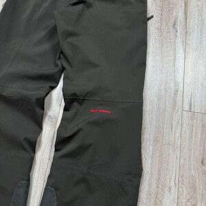 May include: Black outdoor pants with the red "Mammut" logo on the leg. The pants appear to be made of a durable, water-resistant material. The pants are folded and lying on a light-colored wooden surface.
