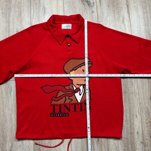 Pu&ograve; includere: Una polo rossa a maniche lunghe con un'illustrazione a cartoni animati di Tintin, un reporter, con cappello e cravatta marroni. La maglia ha un colletto, una chiusura con bottoni e la scritta "TINTIN REPORTER" in basso.