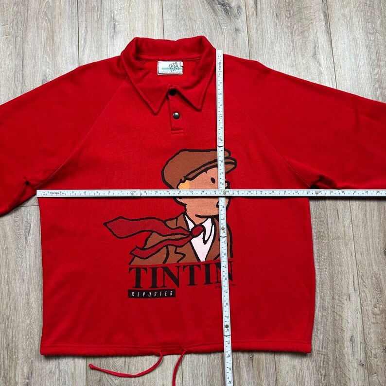 Pu&ograve; includere: Una polo rossa a maniche lunghe con un'illustrazione a cartoni animati di Tintin, un reporter, con cappello e cravatta marroni. La maglia ha un colletto, una chiusura con bottoni e la scritta "TINTIN REPORTER" in basso.