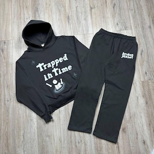 Może przedstawiać: Czarny zestaw bluzy z kapturem i pasujących spodni dresowych. Bluza z kapturem ma napis "Trapped in Time" z grafiką. Spodnie dresowe mają nadruk "broken planet" na nogawce. Zestaw jest wykonany z miękkiego materiału.