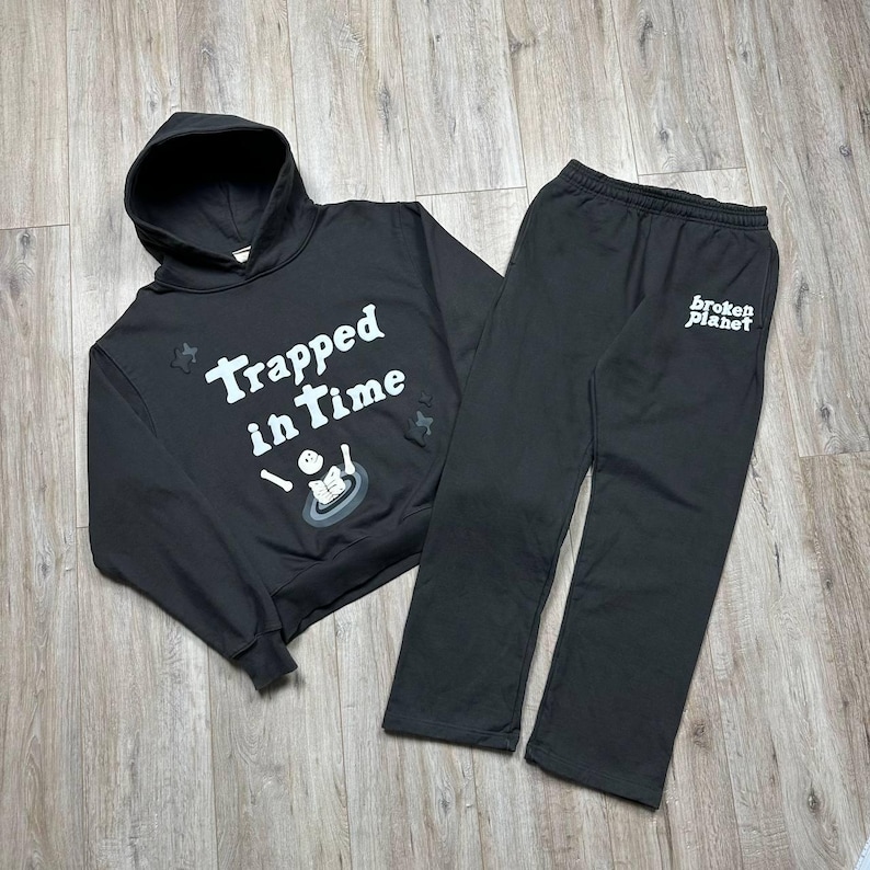 Może przedstawiać: Czarny zestaw bluzy z kapturem i pasujących spodni dresowych. Bluza z kapturem ma napis "Trapped in Time" z grafiką. Spodnie dresowe mają nadruk "broken planet" na nogawce. Zestaw jest wykonany z miękkiego materiału.