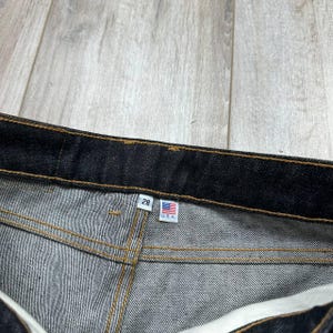 May include: Close-up of dark denim jeans with visible stitching. A tag displays the number "28" and a small American flag with "U.S.A." text. The jeans are on a light wood-grain surface.