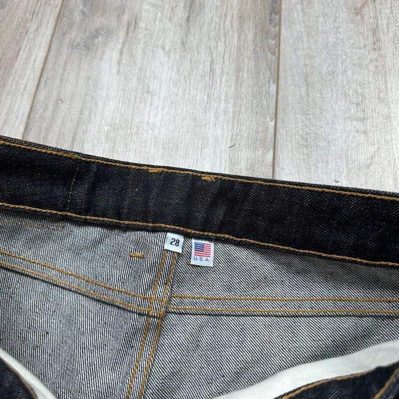 May include: Close-up of dark denim jeans with visible stitching. A tag displays the number "28" and a small American flag with "U.S.A." text. The jeans are on a light wood-grain surface.