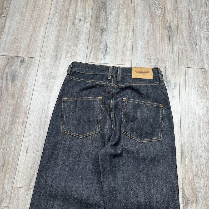 May include: Dark wash denim jeans with a straight leg cut. The jeans feature two back pockets with contrasting stitching and a leather patch on the waistband. The jeans are displayed on a light wood grain surface.