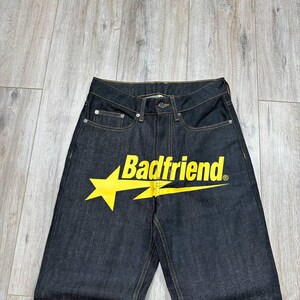 May include: Dark wash denim jeans featuring the text "Badfriend" in yellow with a yellow starburst design. The jeans have classic five-pocket styling and a button and zipper closure.