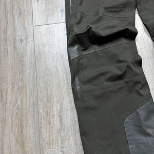 May include: Dark olive-green outdoor pants with reinforced knee and lower leg panels. The word "NIRVANA" is printed in gray on the side of the pants. The pants are made of a durable, water-resistant material.