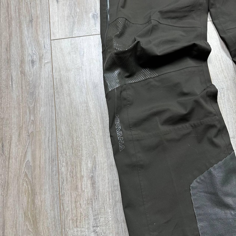 May include: Dark olive-green outdoor pants with reinforced knee and lower leg panels. The word "NIRVANA" is printed in gray on the side of the pants. The pants are made of a durable, water-resistant material.