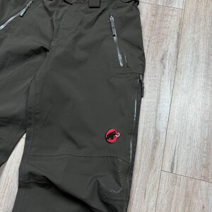 May include: Dark olive-green outdoor pants with multiple zippered pockets and a red logo patch. The pants have a reinforced knee area and a zippered fly. The pants are designed for outdoor activities.