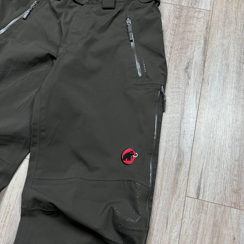 May include: Dark olive-green outdoor pants with multiple zippered pockets and a red logo patch. The pants have a reinforced knee area and a zippered fly. The pants are designed for outdoor activities.