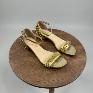May include: A pair of metallic gold sandals featuring an ankle strap with a buckle closure, a low block heel, and a front strap. The heel and front strap are adorned with numerous sharp, pointed gold spikes. The insole is beige with a visible logo.