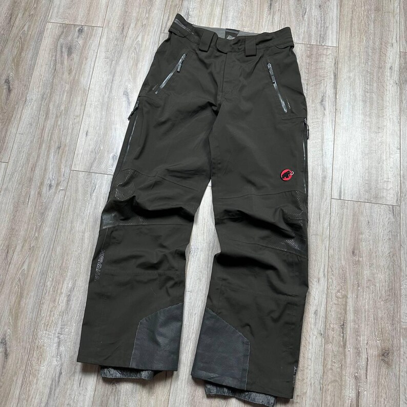 May include: Dark olive green snow pants with multiple zippered pockets and reinforced patches at the knees and ankles. The pants feature a red logo on the right leg and a built-in belt. The pants are designed for winter sports.