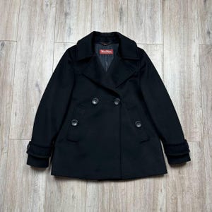 Puede incluir: Un abrigo de lana negro de doble botonadura con cuello con muescas, dos filas de botones y dos bolsillos delanteros. El abrigo tiene un diseño clásico de abrigo marinero y presenta una etiqueta Max Mara. El abrigo se muestra sobre una superficie de madera.