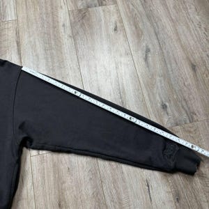 Może przedstawiać: Czarna bluza z długim rękawem z linijką mierzącą długość rękawa. Rękaw ma około 71 cm długości. Na mankiecie wyhaftowano napis "Brixton".