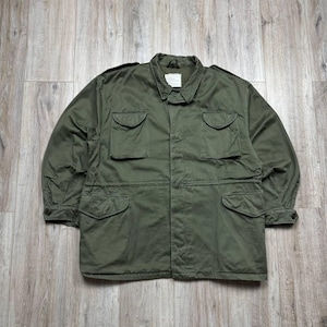 May include: An olive green field jacket featuring a fold-down collar, long sleeves with buttoned cuffs, and four front pockets with buttoned flaps. The garment includes shoulder epaulets and a central front closure, with a white label at the neck.