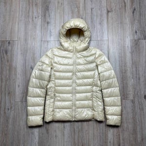 May include: A shiny, pale cream-coloured puffer jacket with a hood and a full-length front zip. The jacket features horizontal quilted stitching throughout the body and sleeves, creating a series of padded sections for a structured appearance.