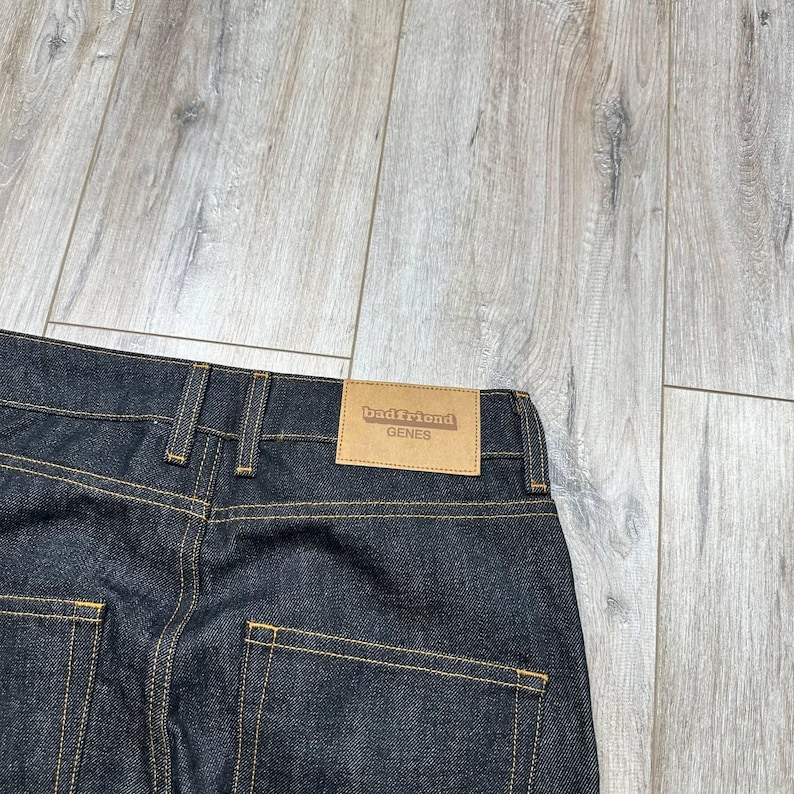 May include: Dark wash denim jeans with visible stitching and a brown leather label. The label reads "badfriend GENES". The jeans are folded, showcasing the back pockets and waistband.
