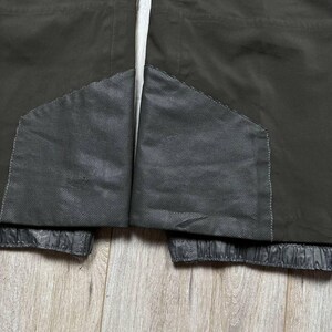 May include: Close-up of a dark olive-green jacket. The jacket features two dark gray mesh panels and a white drawstring. The bottom hem is elasticized. The jacket is made of a durable, water-resistant material.