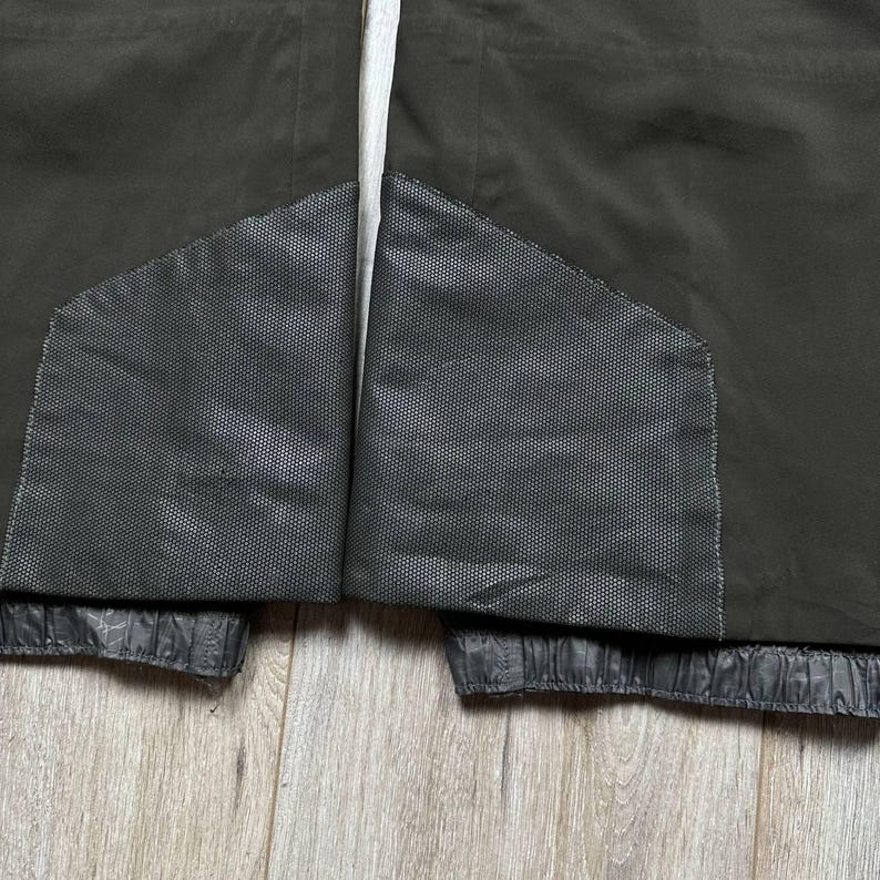 May include: Close-up of a dark olive-green jacket. The jacket features two dark gray mesh panels and a white drawstring. The bottom hem is elasticized. The jacket is made of a durable, water-resistant material.