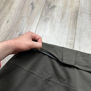May include: Close-up of a dark olive-green garment with a zipper. The garment has a subtle sheen and a small, embossed logo on a flap. The zipper is silver-colored. The fabric appears to be a lightweight, possibly water-resistant material.