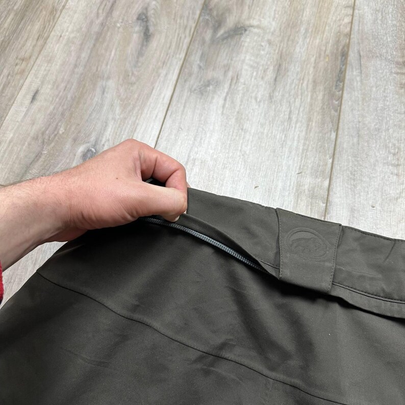 May include: Close-up of a dark olive-green garment with a zipper. The garment has a subtle sheen and a small, embossed logo on a flap. The zipper is silver-colored. The fabric appears to be a lightweight, possibly water-resistant material.