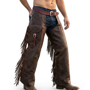 May include: Brown leather chaps with fringe detailing and a pocket. The chaps are worn over blue jeans and feature a decorative belt. The design includes red accents and metal hardware, showcasing a Western style.
